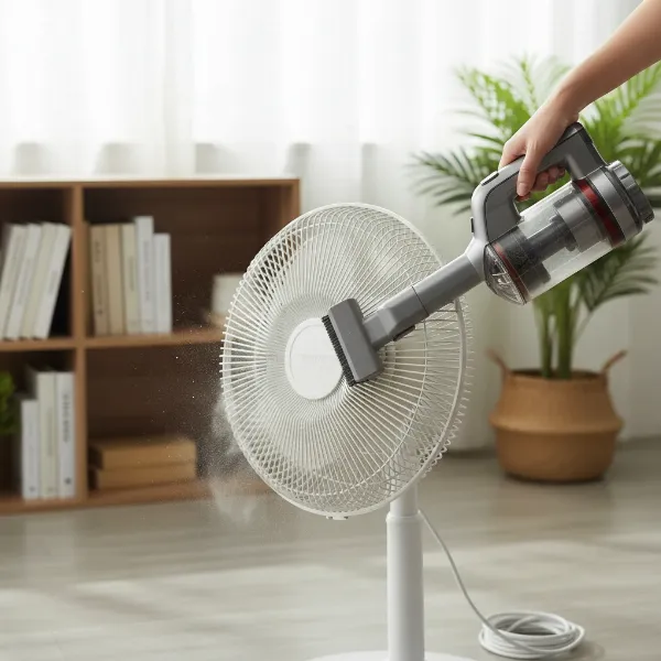 A person is using a vacuum cleaner brush attachment to clean the dusty grill of an electric fan.